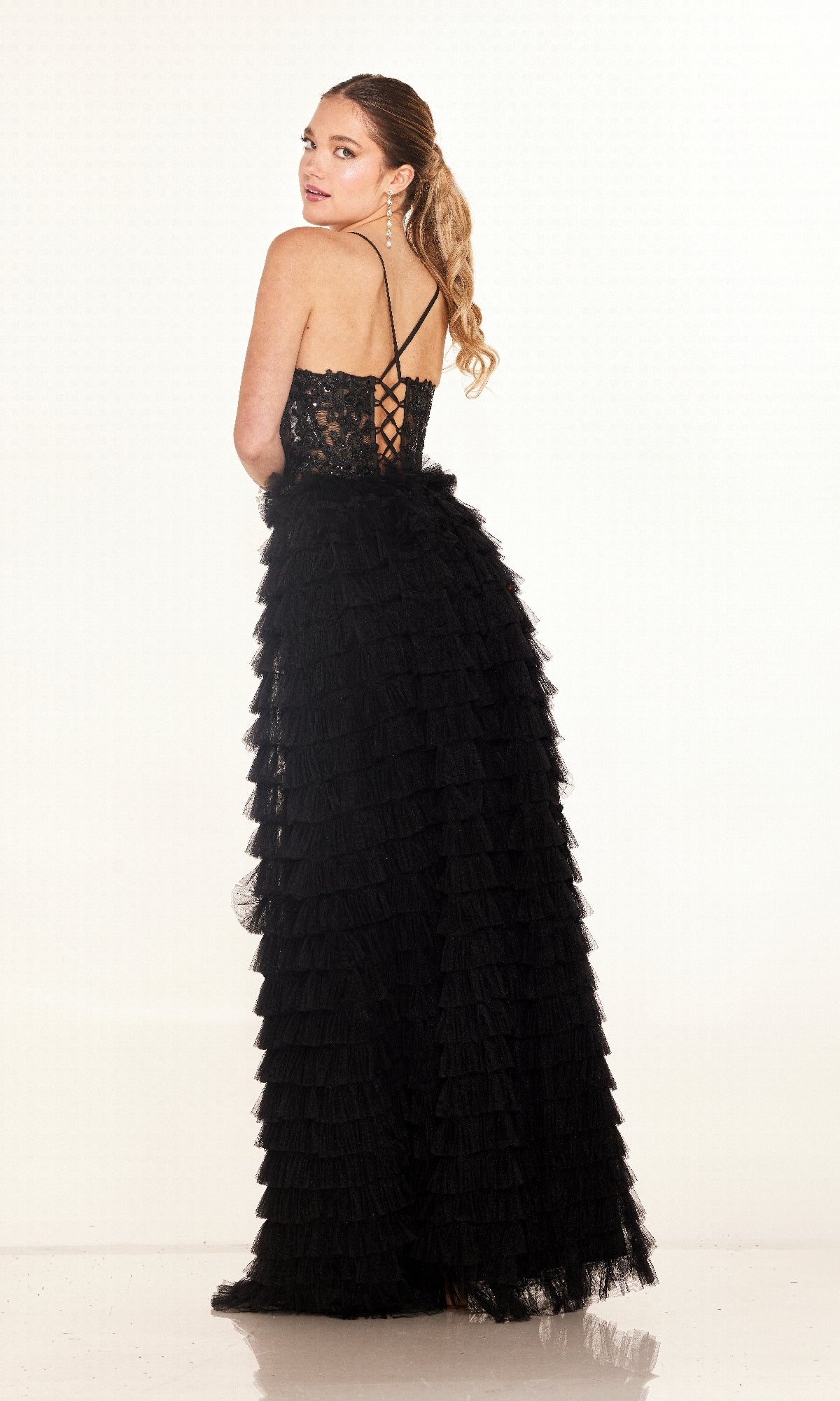 A woman stands looking over her shoulder in the Long Prom Dress: Cecilia Couture 2624, a black evening gown with ruffles, a sheer corset bodice, lace detailing, crisscross straps, and a lace-up back.